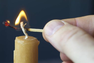 Yakından erkek eli yanan kibrit tutuyor ve koyu arkaplanda sarı mum yakıyor, seçici odaklı.