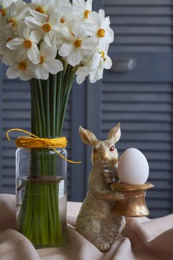 Bahar kompozisyonu, yumurta ve cam vazolu tavşan heykeli bej masa örtüsü üzerine nergis, paskalya konsepti, seçici odak