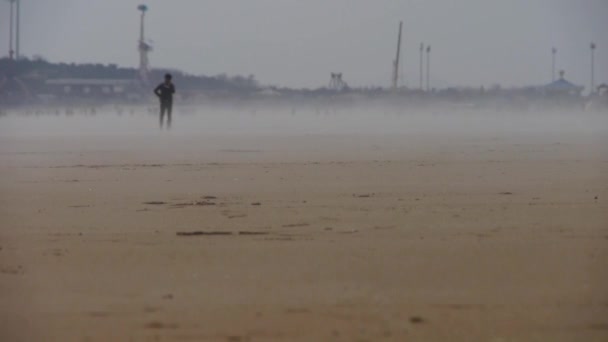 Wiatr wiejący mgłą nad plażą, ludzie chodzący po plaży przeciwko mirażom. — Wideo stockowe
