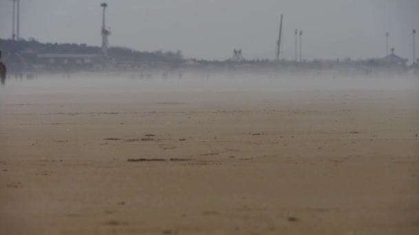 Wiatr wiejący mgłą nad plażą, ludzie chodzący po plaży przeciwko mirażom. — Wideo stockowe