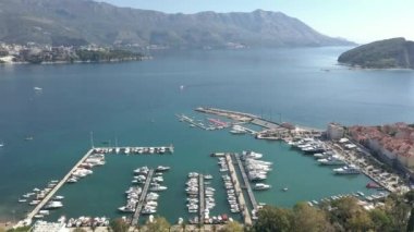 Güneşli bir yaz gününde, Karadağ 'ın Budva kentindeki bir marinanın demirli tekne ve yatlarla çekilmiş hava görüntüleri. Denizin suları zümrüt, arka planda dağlar görünür.