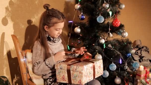 Ein kleines Mädchen in der Nähe des Weihnachtsbaums öffnet ein Neujahrsgeschenk — Stockvideo