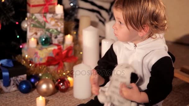 Маленькая девочка играет рядом с елкой с новогодним подарком — стоковое видео