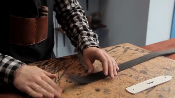 Con un coltello industriale, il maestro taglia la pelle in eccesso sul pezzo sotto la cinghia. Procedura per la fabbricazione di cinture di cuoio — Video Stock