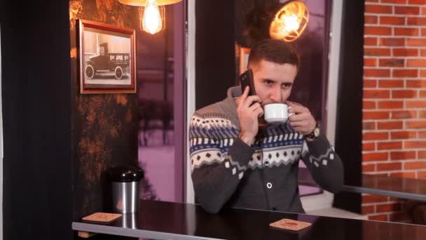 Carismático joven bebe café y se comunica por teléfono en un restaurante — Vídeo de stock