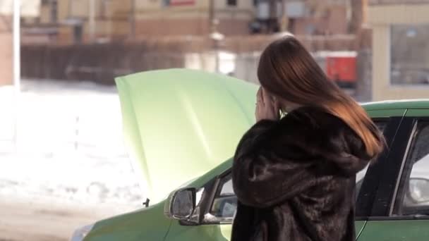 Το χειμώνα, μια έγκυος κορίτσι, στέκεται κοντά ένα ελαττωματικό αυτοκίνητο με μια ανοικτή κουκούλα, προσπαθεί να καλέσετε έναν γερανό με ένα κινητό τηλέφωνο — Αρχείο Βίντεο