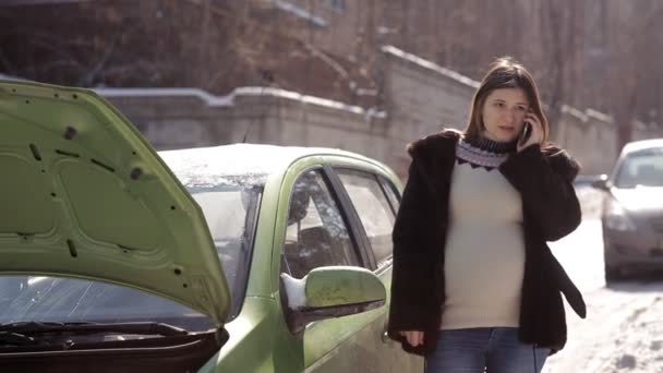 Зимой беременная девушка, стоящая возле неисправной машины с открытым капотом, пытается вызвать эвакуатор с мобильного телефона — стоковое видео