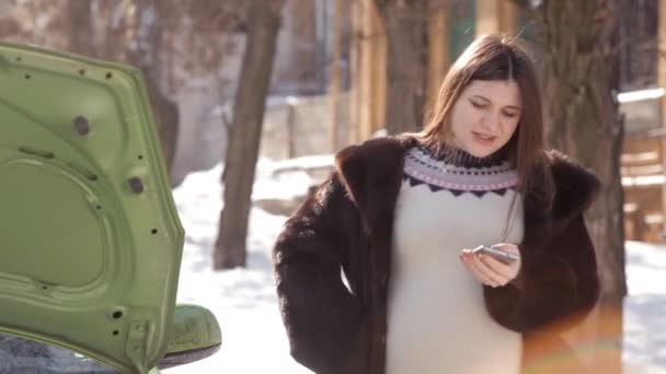 In inverno, una ragazza incinta, in piedi vicino a una macchina difettosa con un cappuccio aperto, cercando di chiamare un carro attrezzi con un telefono cellulare — Video Stock