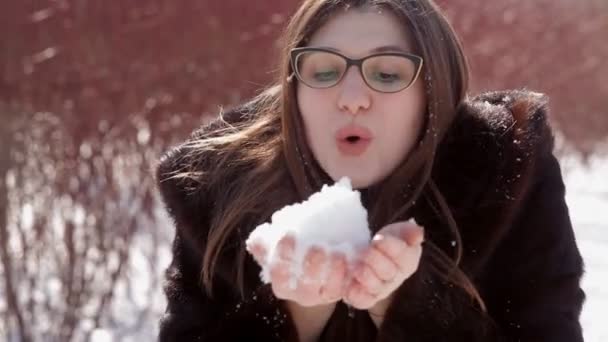 Glad tjej med glasögon och mink päls blåses upp snön från händerna — Stockvideo