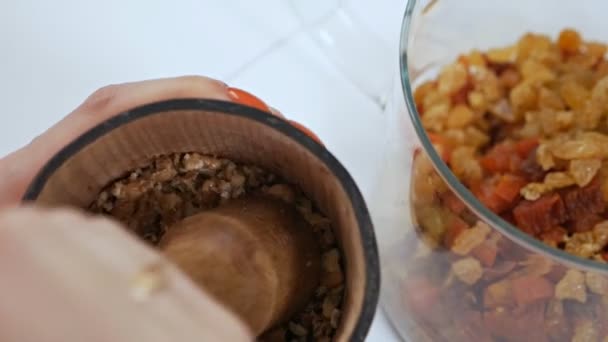 Woman chopping walnuts in a wooden mortar and puts them in a glass bowl with dried fruits — Stock Video