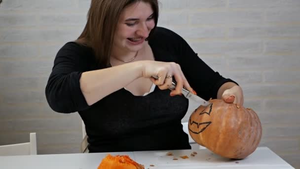 Mulher com um olhar louco e olhar, preparando uma abóbora em uma festa de Halloween na mesa da cozinha, uma menina corta a boca com uma faca — Vídeo de Stock