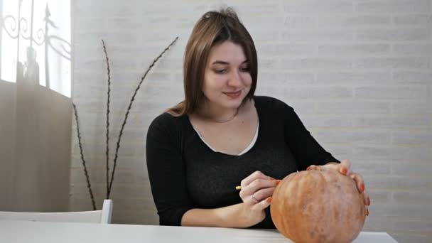 Concetto di Halloween, ragazza felice seduta a un tavolo con zucche, occhi e bocca dipinti su una zucca di Halloween — Video Stock