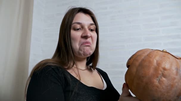 Uma jovem segura uma abóbora desaparecida em suas mãos e enruga seu rosto, faz várias expressões faciais. Imagem de abóbora — Vídeo de Stock