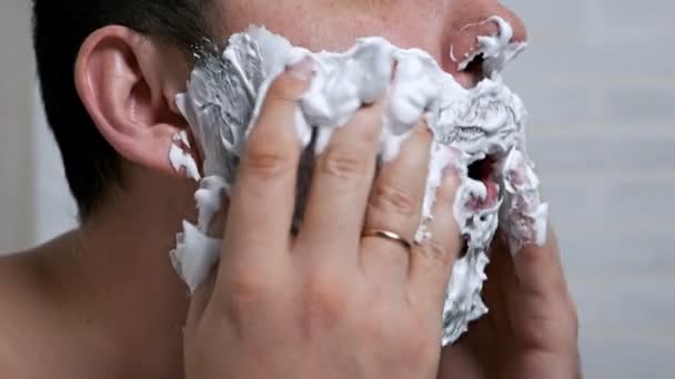 Macho guapo causa espuma de afeitar descuidado en su cara y sonríe extrañamente al mismo tiempo.Depilación en la cara — Vídeo de stock