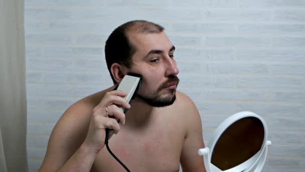 Bir adam sakalını ve bıyığını evde elektrikli jiletle tıraş eder, aynanın önünde saç kesilir. Erkekler umursar — Stok video