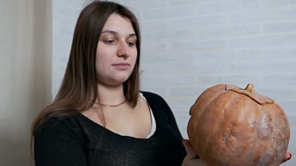 Una joven sostiene una calabaza perdida en sus manos y arruga su cara, hace varias expresiones faciales. Imagen de calabaza — Vídeos de Stock