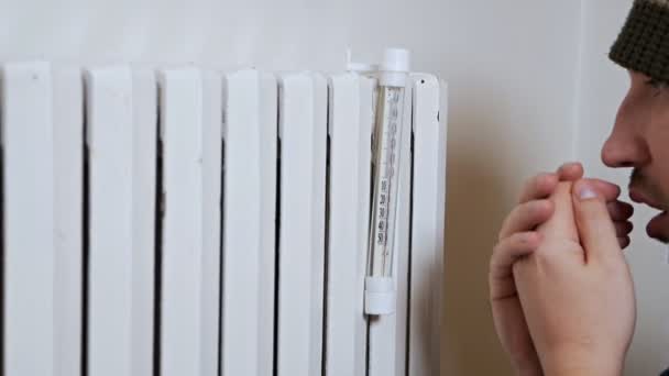 Un uomo misura la temperatura dell'aria in una casa in un giorno d'inverno vicino a una batteria. Un riscaldamento debole e scadente. In abiti caldi in casa, appartamento — Video Stock