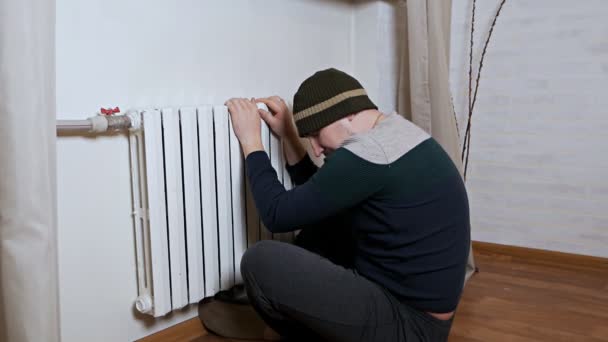 Um homem de chapéu aquece as mãos num radiador de aquecimento perto da parede. Frio no apartamento, sistema de aquecimento pobre — Vídeo de Stock