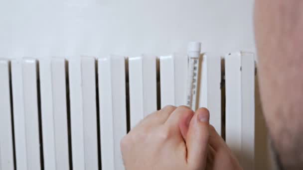 Un hombre mide la temperatura del aire en una casa en un día de invierno cerca de una batería. Débil, pobre calefacción. En ropa de abrigo en la casa, apartamento — Vídeos de Stock