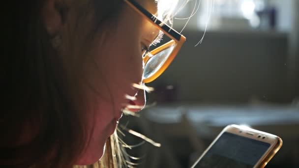 Вид сбоку на молодую женщину в очках, смотрящую на смартфон возле окна — стоковое видео