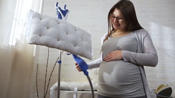 Hermosa mujer embarazada joven en traje gris sobre fondo blanco utilizando el sistema de vapor para planchar almohadas en casa — Vídeos de Stock