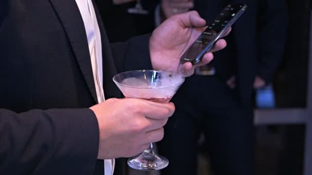 Um homem apresentável em um terno clássico segura um coquetel na mão e usa um telefone celular em uma festa. Bebida alcoólica criativa — Vídeo de Stock