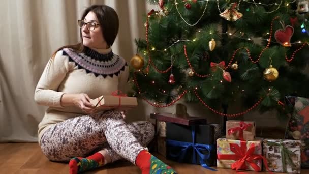 Una jovencita abre un regalo de Navidad sentada en el suelo cerca de un árbol de Navidad mientras nadie la ve — Vídeos de Stock