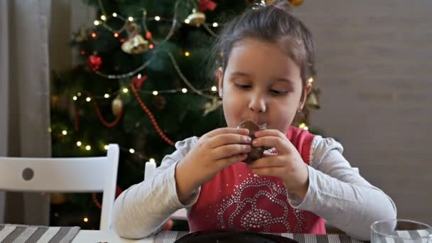 Chica sentada junto al árbol de Navidad comiendo chocolate Santa Claus en Nochebuena. Celebrando la Navidad en casa — Vídeos de Stock