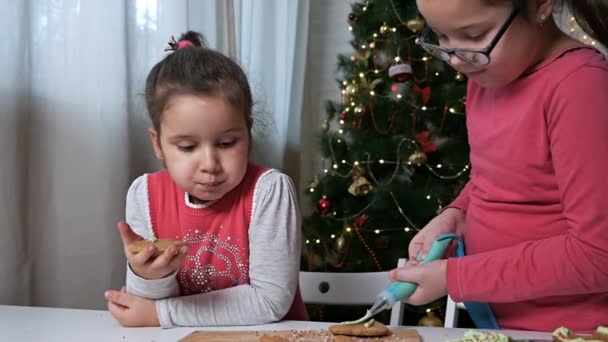 Két aranyos kisgyerek, lányok-nővérek, készítsenek mézeskalácsot karácsonyra egy gyönyörű karácsonyfa hátterében. Házi főzés. Finom mézeskalács, enni és főzni — Stock videók