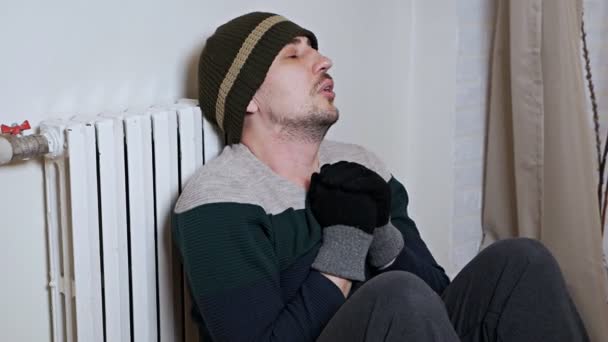 Ένας άντρας στο σπίτι με καπέλο και ζεστά ρούχα απολαμβάνει ζεστές μπαταρίες. Παγετός κρύου χειμώνα — Αρχείο Βίντεο