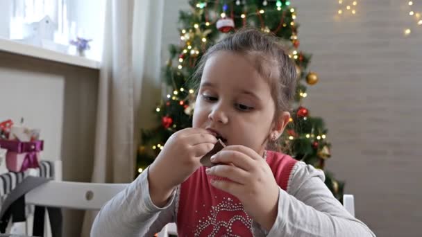 Flicka sitter vid granen äter choklad jultomten på julafton. Fira jul hemma — Stockvideo