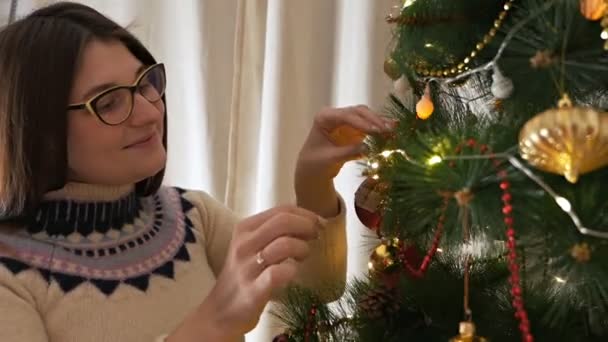 Jong gelukkig vrouw in kerst trui glimlachen en versieren kerstboom hang speelgoed — Stockvideo