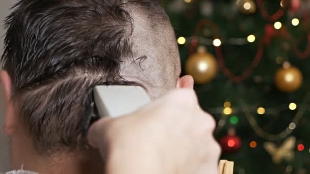 Corte de pelo para hombre con afeitadora eléctrica. Peluquería peinado en casa. Las manos femeninas usan una máquina de afeitar eléctrica para quitar el pelo de la cabeza de un hombre contra el fondo de un árbol de Navidad — Vídeos de Stock