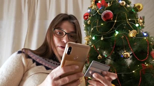 Uma jovem perto da árvore de Natal, com um telefone, pede presentes para o Ano Novo, pagando com um cartão de crédito. Despesas de Ano Novo — Vídeo de Stock