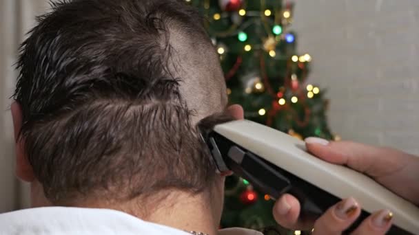 Corte de pelo para hombre con afeitadora eléctrica. Peluquería peinado en casa. Las manos femeninas usan una máquina de afeitar eléctrica para quitar el pelo de la cabeza de un hombre contra el fondo de un árbol de Navidad — Vídeo de stock