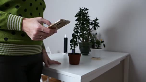 Het lichaam, de figuur van een jong meisje op de voorgrond, op de achtergrond planten in potten. In de hand van het meisje, aan de telefoon, waarop ze foto 's bekijkt van huisplanten aan de telefoon. eco blogger — Stockvideo