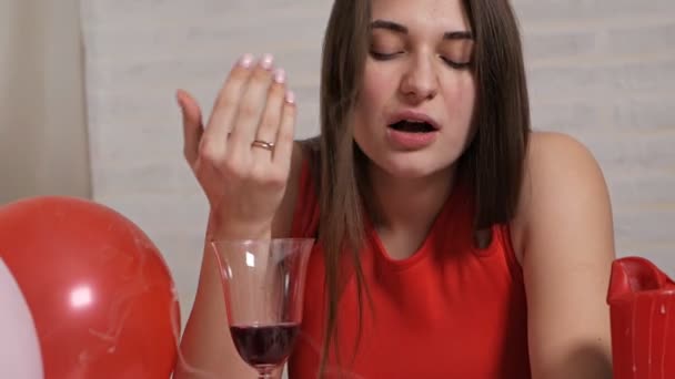 Een mooi meisje in een rode jurk alleen aan tafel drinkt wijn uit een glas terwijl ze dronken is. Eenzame Valentijn — Stockvideo