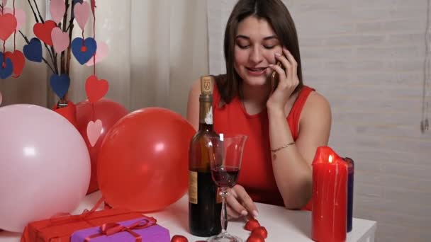 Noite romântica com velas e vinho, uma menina de vestidos vermelhos sentada em uma mesa com um telefone e esperando por seu amado, Dia dos Namorados — Vídeo de Stock