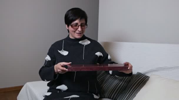 Eine ältere Frau freute sich über ein Geschenk. glückliche ältere Frau mit einer Schachtel auf einem Sofa im Wohnzimmer sitzend — Stockvideo