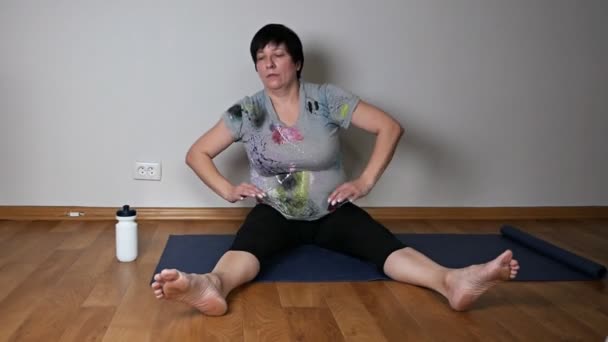 Активная положительная пожилая женщина делает разминку, растягиваясь сидя на полу в гостиной — стоковое видео