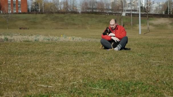 Un homme s'assoit sur l'herbe et étreint son chien berger allemand sur une pelouse herbeuse. Mouvement lent — Video