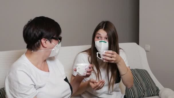 Een jonge vrouw en haar moeder drinken koffie in een medisch masker, zittend op een bank thuis tijdens de quarantaine. Sociale afstand. Virussen — Stockvideo