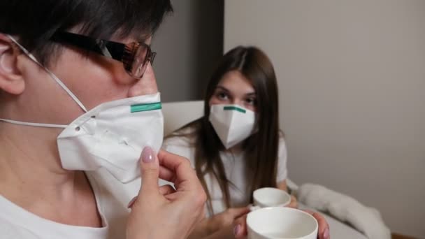 Een jonge vrouw en haar moeder drinken koffie in een medisch masker, zittend op een bank thuis tijdens de quarantaine. Sociale afstand. Virussen — Stockvideo