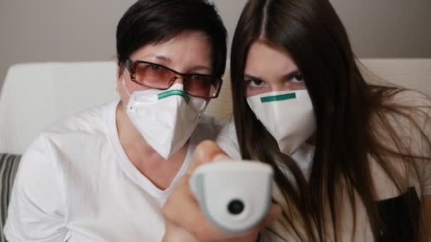 Mother and daughter at home with a protective medical mask use an electronic thermometer to measure temperature. The epidemic of influenza, Covid-19, coronavirus — Stock Video