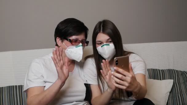 Una madre y su hija llevando una máscara protectora y sentadas en el sofá en casa juntas usando un teléfono móvil. Protección contra el virus Corona, brote de COVID 19. Concepto de vida saludable y segura — Vídeo de stock