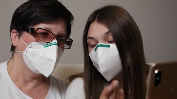 Una madre y su hija llevando una máscara protectora y sentadas en el sofá en casa juntas usando un teléfono móvil. Protección contra el virus Corona, brote de COVID 19. Concepto de vida saludable y segura — Vídeos de Stock