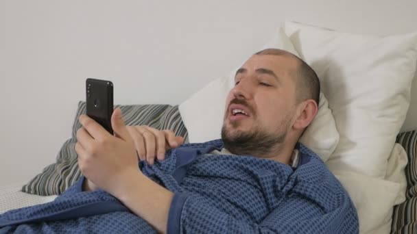 Uomo pigro che utilizza un telefono cellulare sdraiato su un letto a casa — Video Stock
