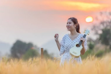 Ukulele tutarak ve gün batımı zamanında yaz aylarında arpa sahasında rahatlatıcı güzel Asyalı kadın