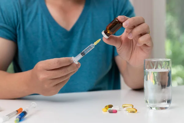 Young man holding injection needle and vaccine in hand with assorted pharmaceutical medicine pills on table