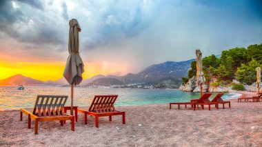 Budva yakınlarındaki Sveti Stefan 'da şezlonglar ve şemsiyelerle plajda dramatik bir gün batımı. Konum: Budva Riviera, Karadağ, Adriyatik denizi, Avrupa
